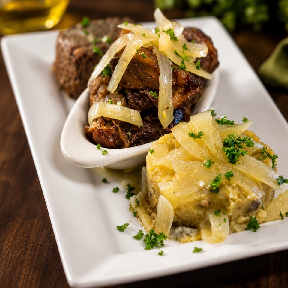 Plated entrée featuring braised meat and a golden vegetable casserole topped with caramelized onions and fresh parsley.