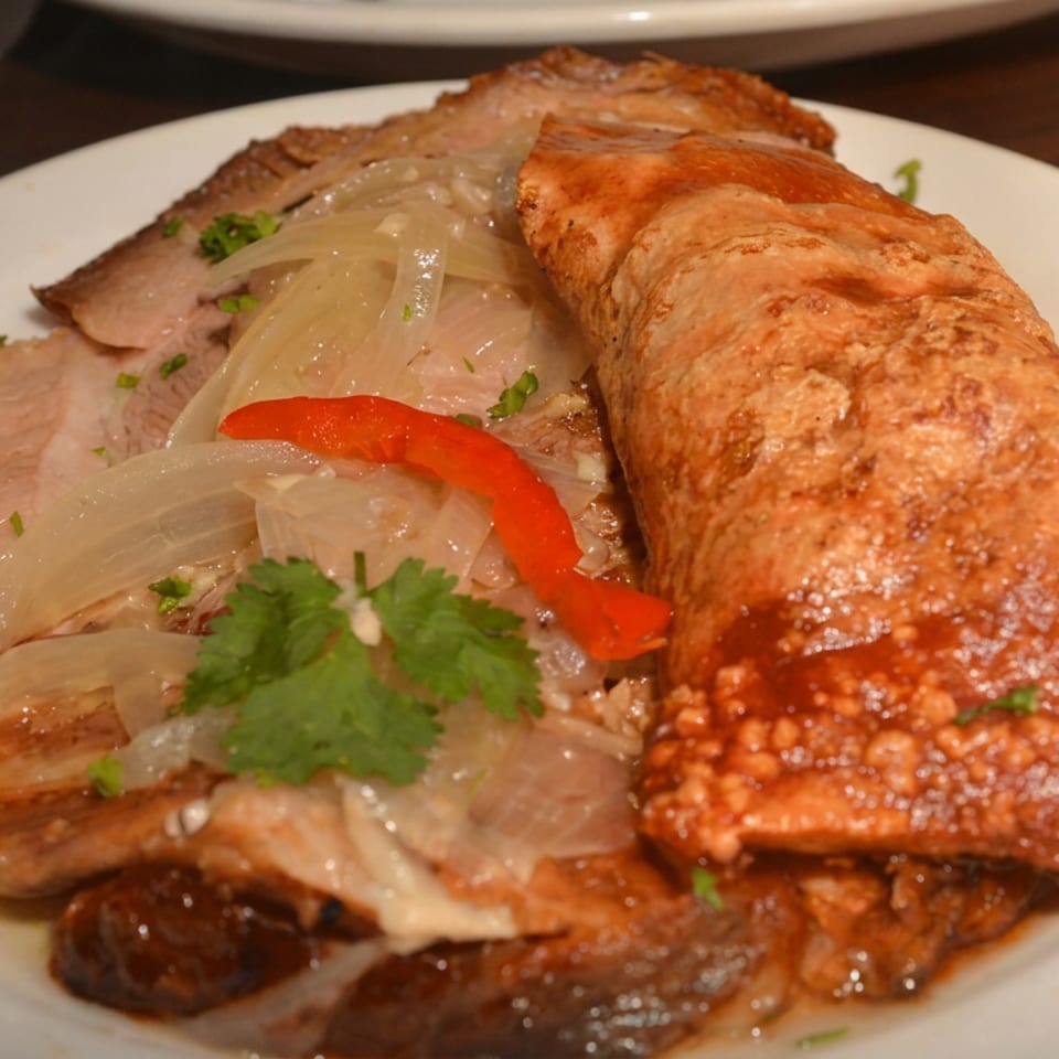 Roasted pork and sausage topped with sautéed onions, red pepper, and cilantro on a white plate.