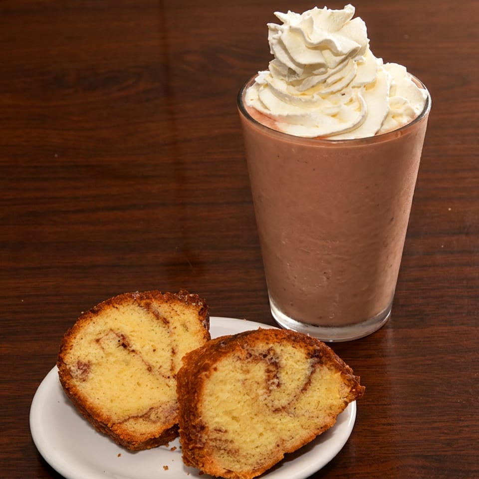 Chocolate milkshake topped with whipped cream beside two cinnamon-swirled cake slices on a white plate atop a wooden table.