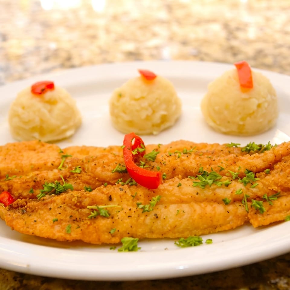 Breaded fried fish fillets with mashed potato portions, lime wedges, red pepper, and chopped herbs on a white plate.