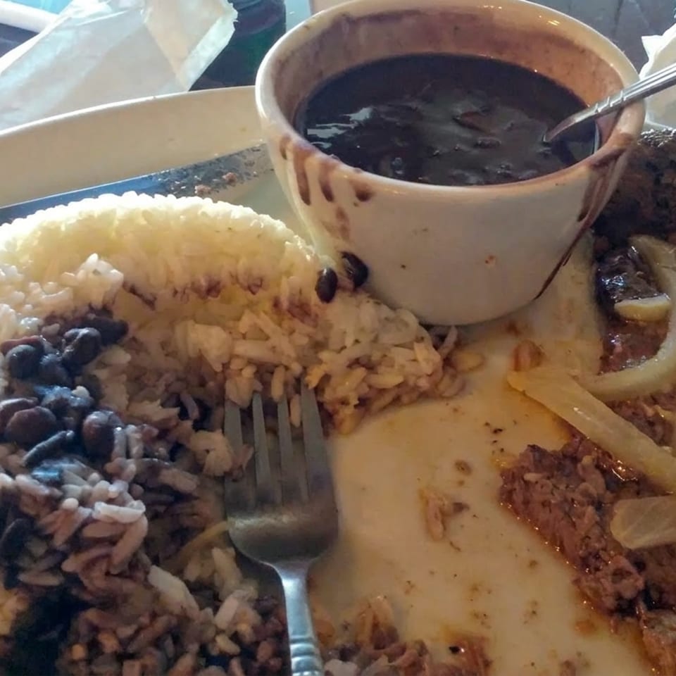 Plate of rice and black beans, grilled steak topped with onions, French fries, and a bowl of dark sauce.