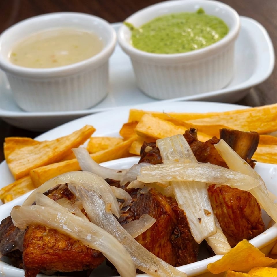 Plate of roasted meat topped with onions, served with golden plantain chips, white sauce, and green dipping sauce.
